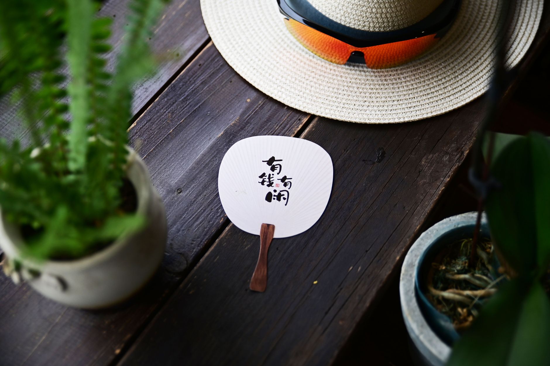 A traditional Chinese fan with text on a rustic wooden table, next to a straw hat and plants.
