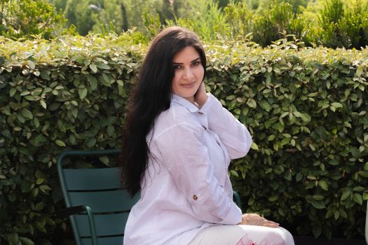 Elegant woman sitting outdoors in front of greenery, wearing a white shirt.
