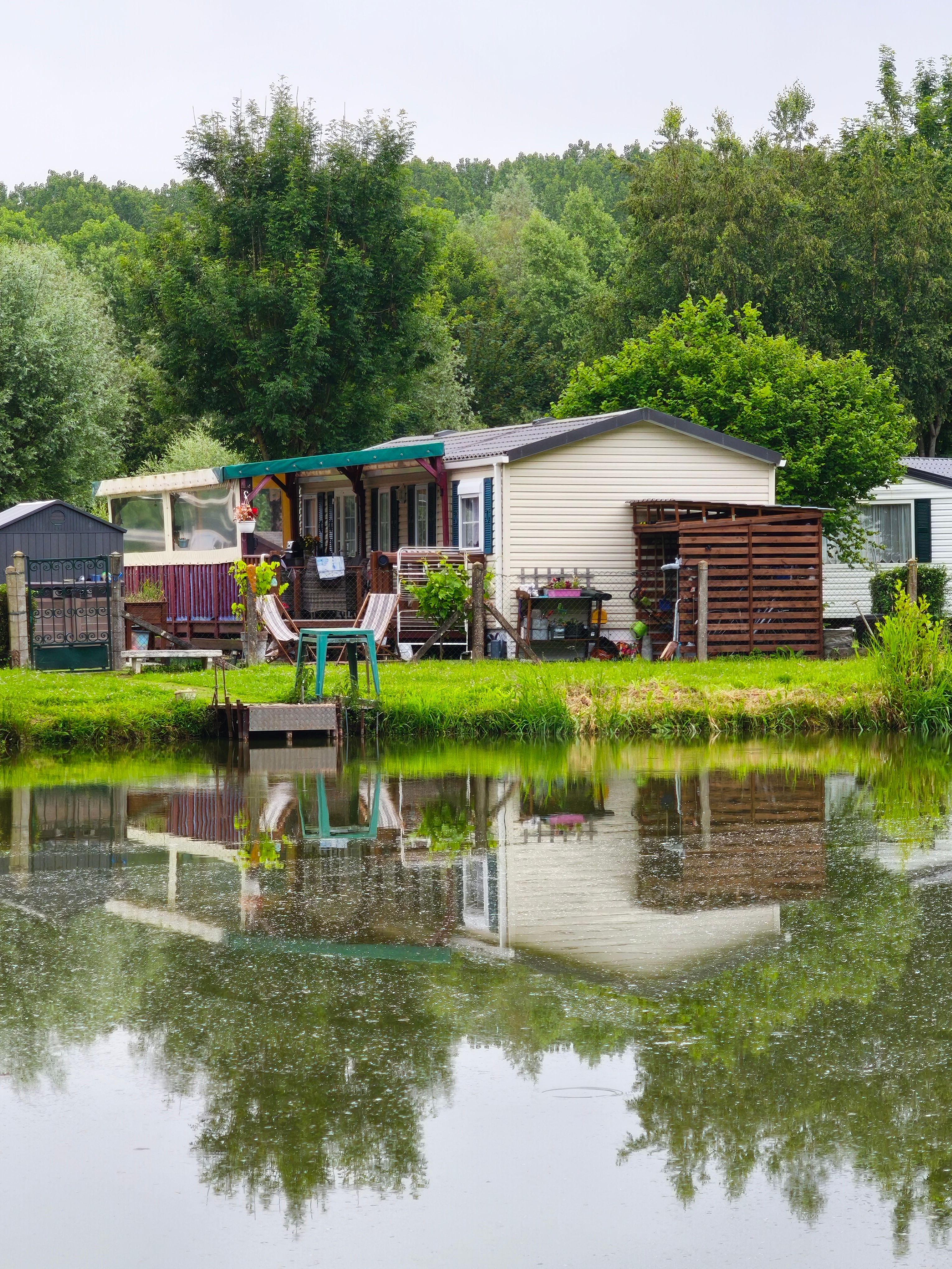 A serene mobile home sits by a tranquil lake surrounded by lush greenery, ideal for nature retreats.