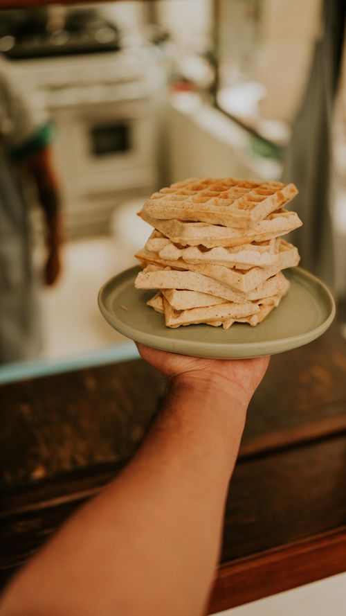 Free A hand holding a stack of freshly made waffles on a green plate in a cozy kitchen setting. Stock Photo