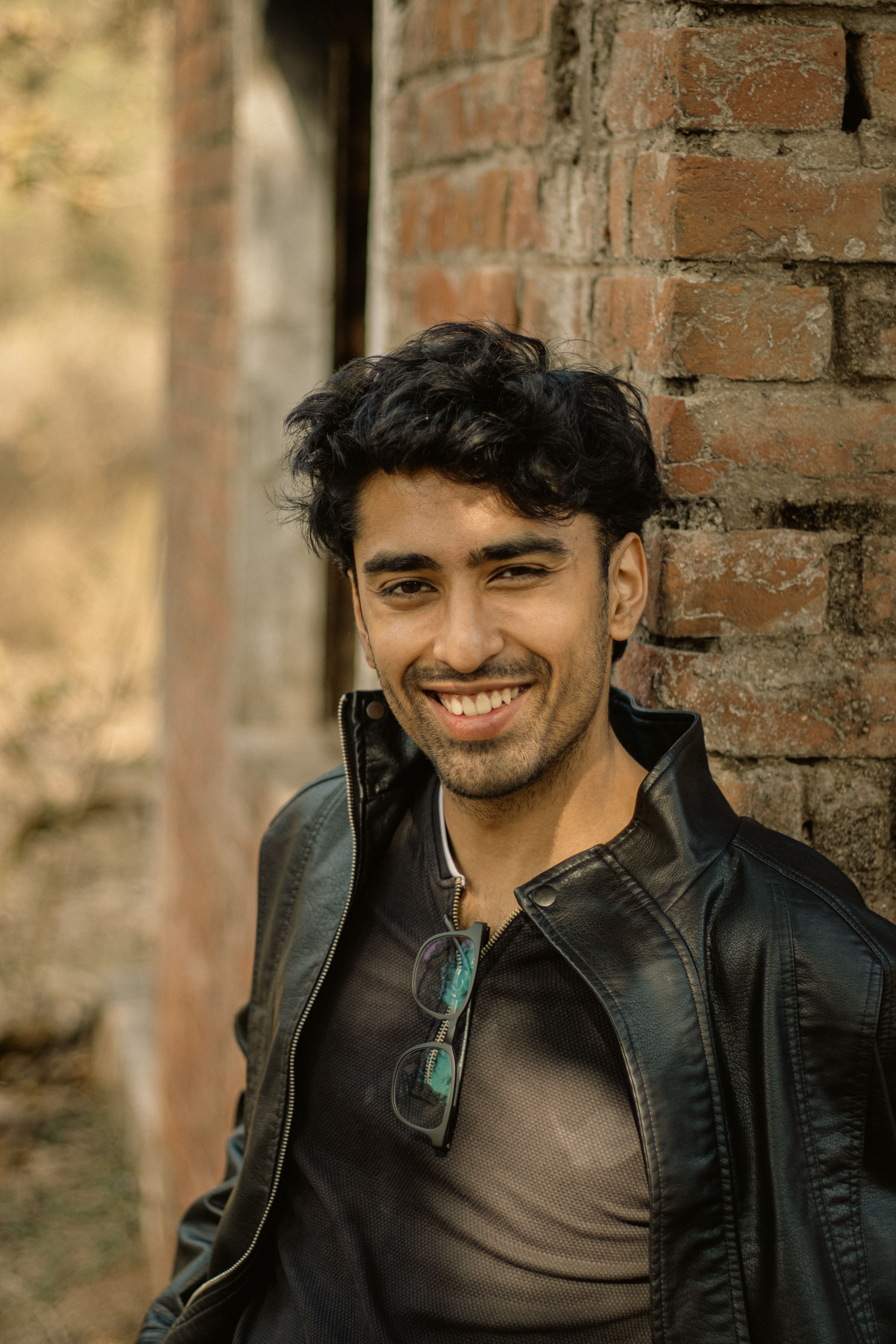 Portrait of a young man in a black leather jacket smiling warmly against a brick wall.