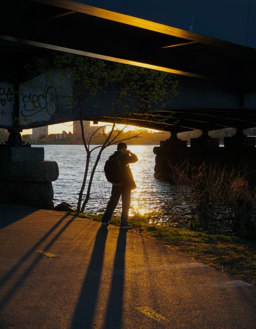 Free A photographer captures the sunset under a bridge, creating a stunning silhouette against the river. Stock Photo