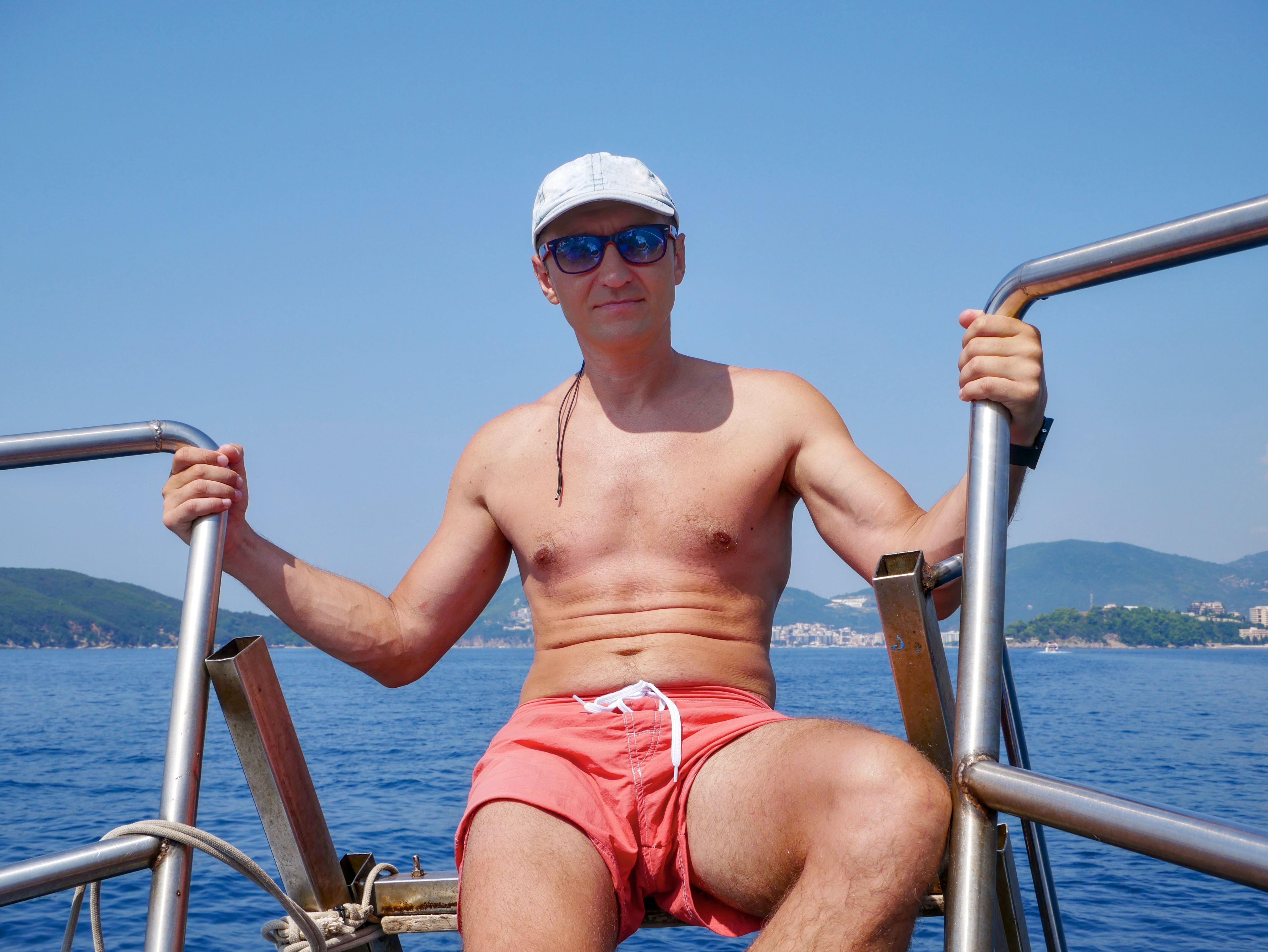Man enjoying a sunny day on a boat, wearing sunglasses and swim trunks against a scenic ocean backdrop.