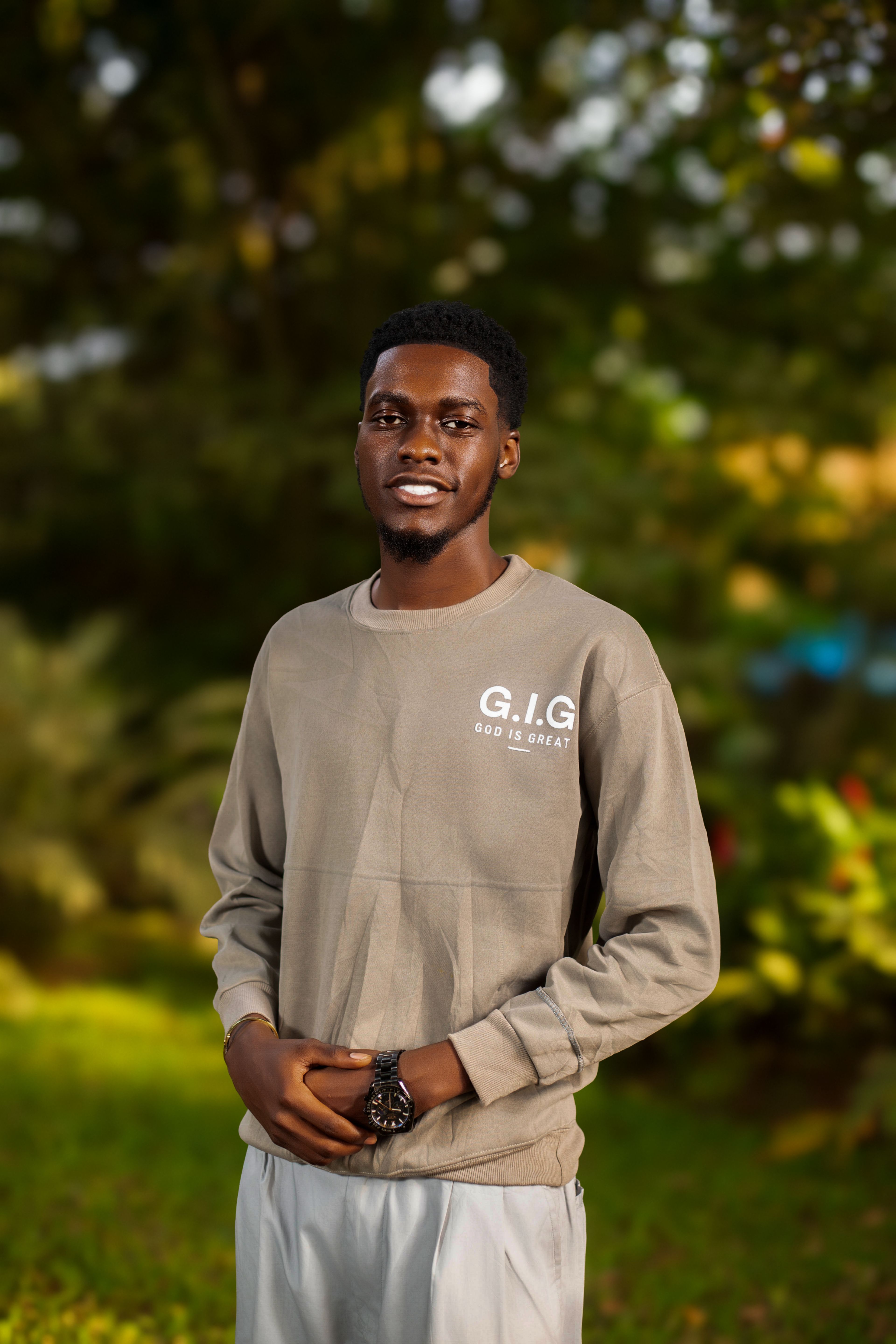 A young man with a relaxed smile stands in a sunny garden wearing a casual sweatshirt.