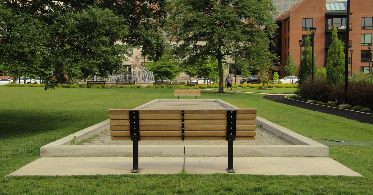 An urban park featuring empty benches, a walking path, lush green grass, and city buildings in the background.