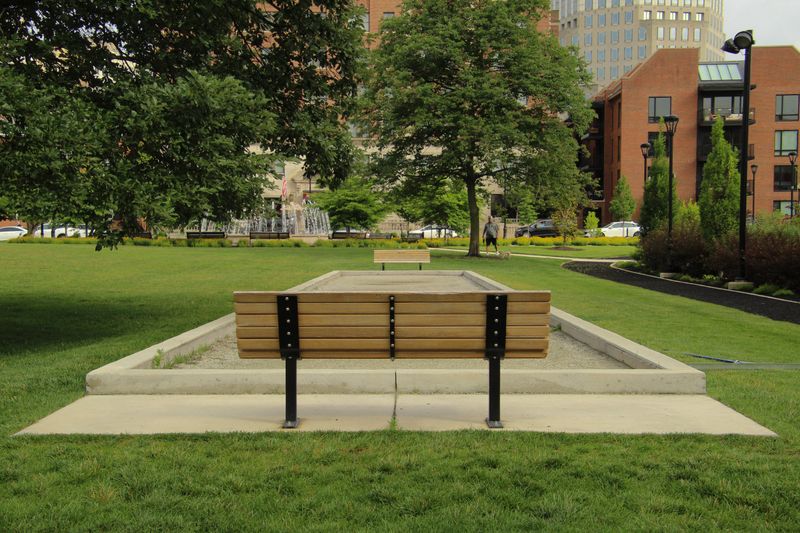 An urban park featuring empty benches, a walking path, lush green grass, and city buildings in the background.