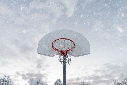 Snow-covered basketball hoop with falling snowflakes under a cloudy winter sky.
