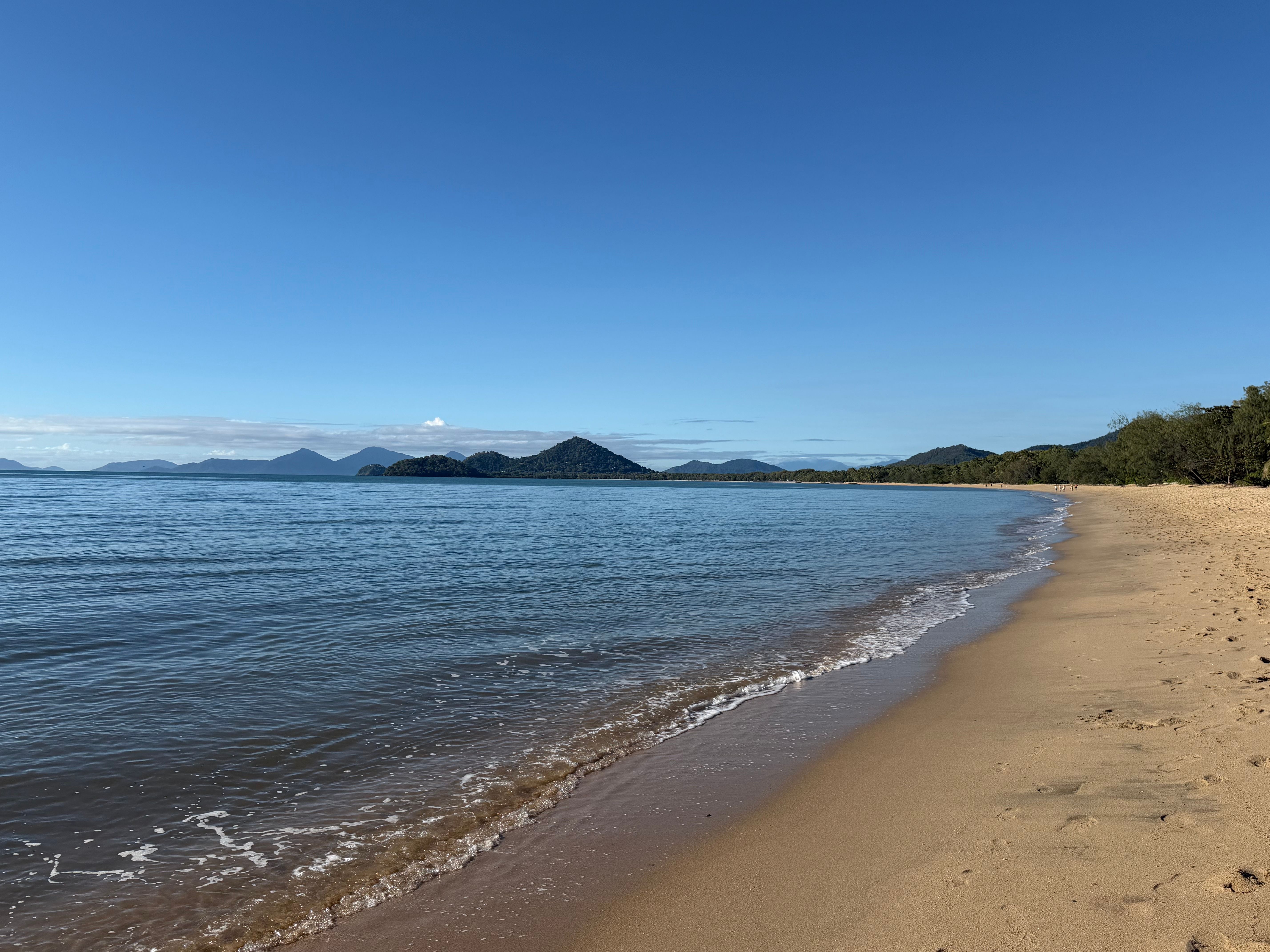 A broad tropical shoreline under clear skies, echoing the quiet scale of North Queensland coastal towns before the internet arrives.
