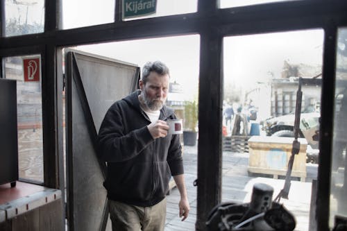 Man in Black Jacket and Brown Pants Standing Beside Glass Door