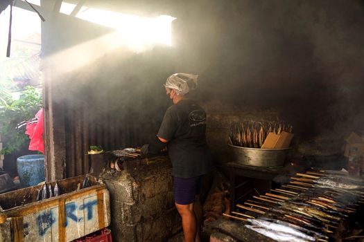 A local Balinese worker grilling fish in a rustic smokehouse, showcasing traditional culinary methods.