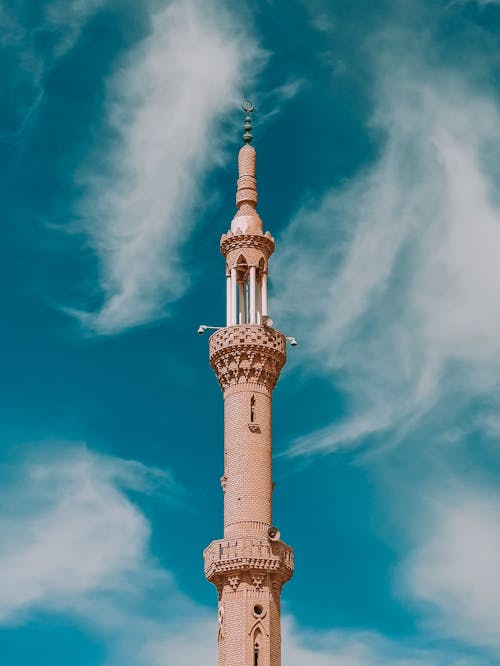 From below scenery view of aged high masonry tower with columns and decorative elements on walls and pointed spire on top with lamps under sky with clouds in daylight