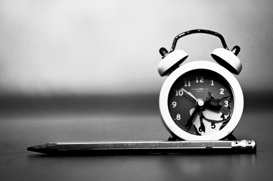Black and white photo of a vintage alarm clock beside a pencil, emphasizing time and writing.