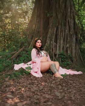 Maternity portrait of a woman in a pink gown sitting under a large tree in a forest.