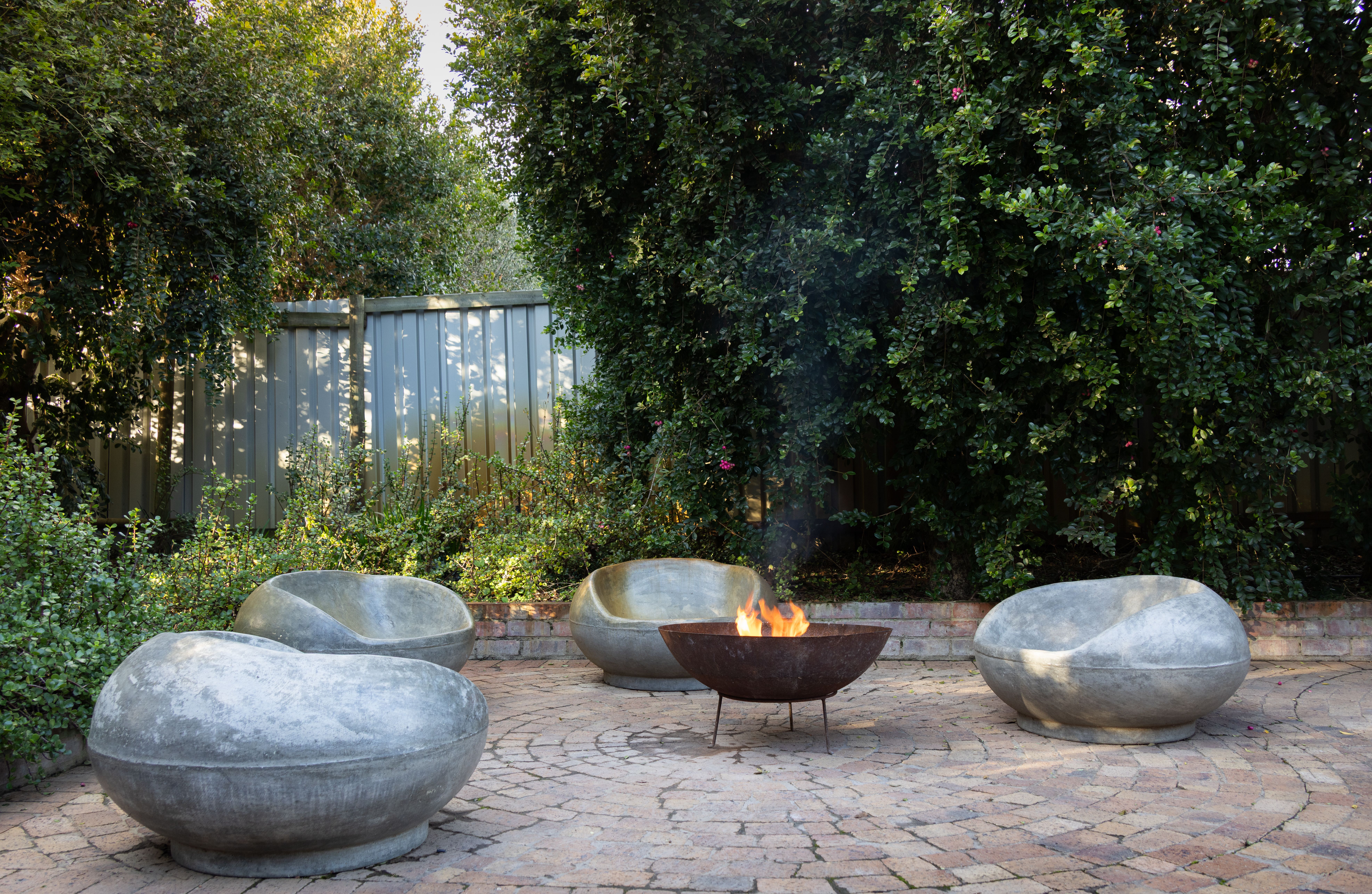 A modern gas fire pit with a sleek stone surround, set into a bluestone patio with lush green plantings.
