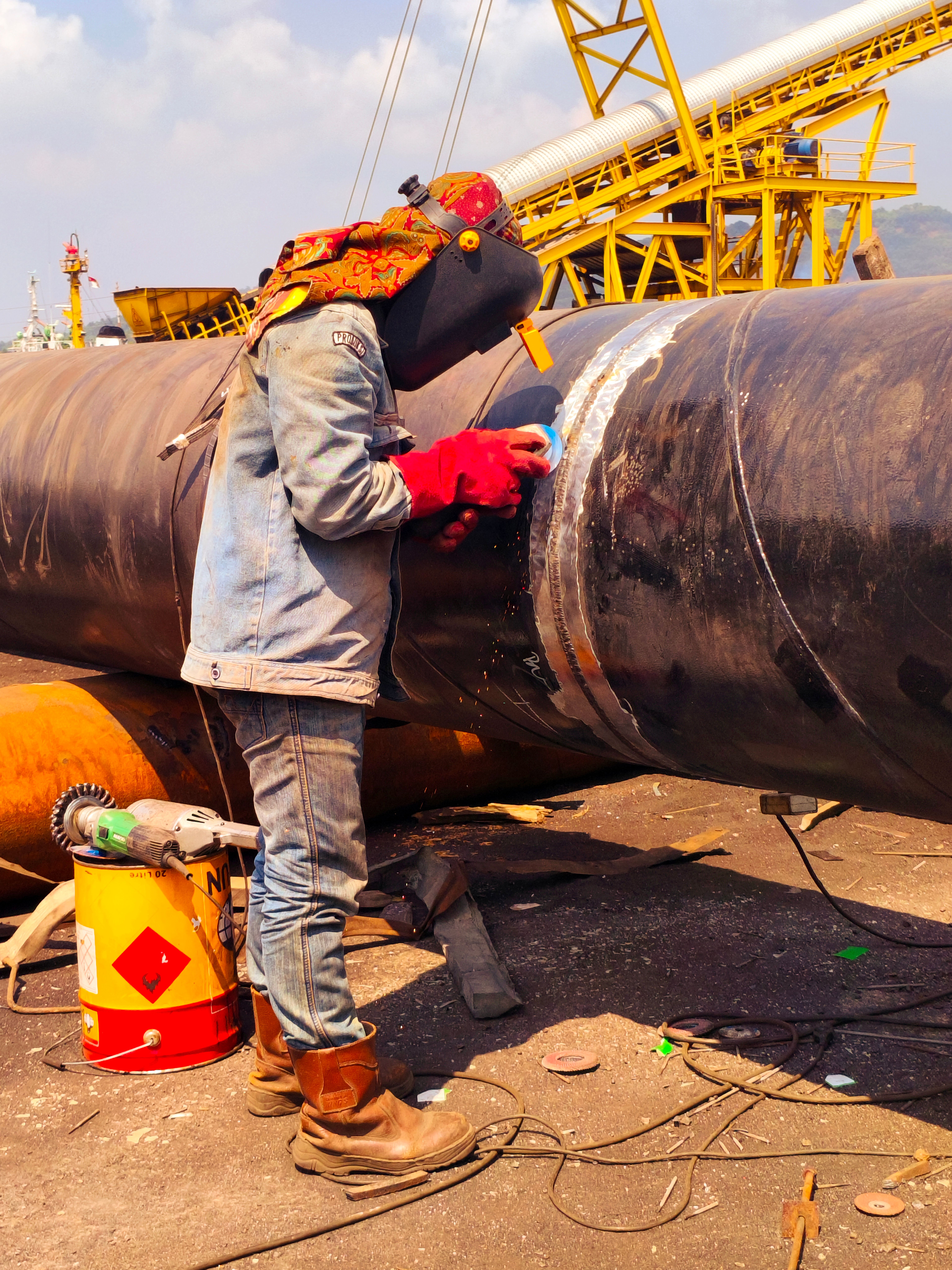 Pipe welder working a joint on large-diameter pipe under a yard crane