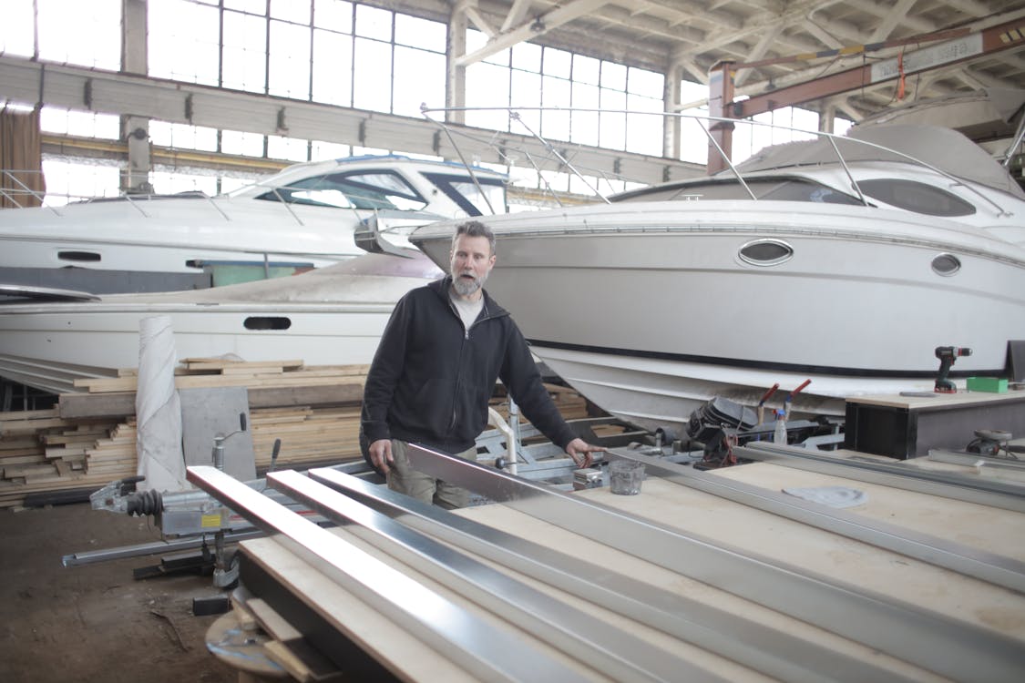Serious brutal male mechanic wearing casual clothes standing near metal details in spacious hangar with yachts