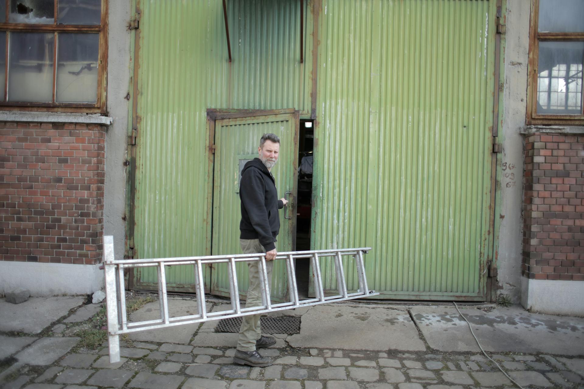 Man holding a stepladder while entering a large green warehouse door.