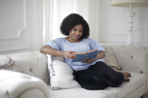 Free A joyful woman with afro hair lounges on a sofa, enjoying a book in a cozy living room. Stock Photo
