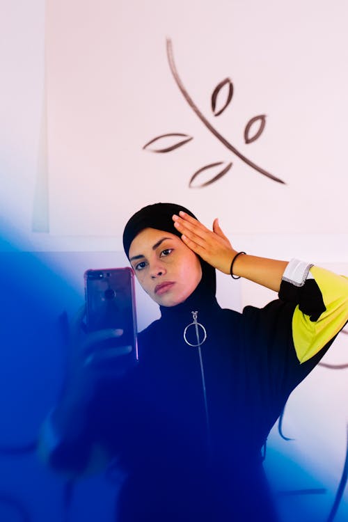 Free Concentrated young ethnic female wearing sportswear with tight hood covering head and taking selfie against white wall with abstract illustrations Stock Photo