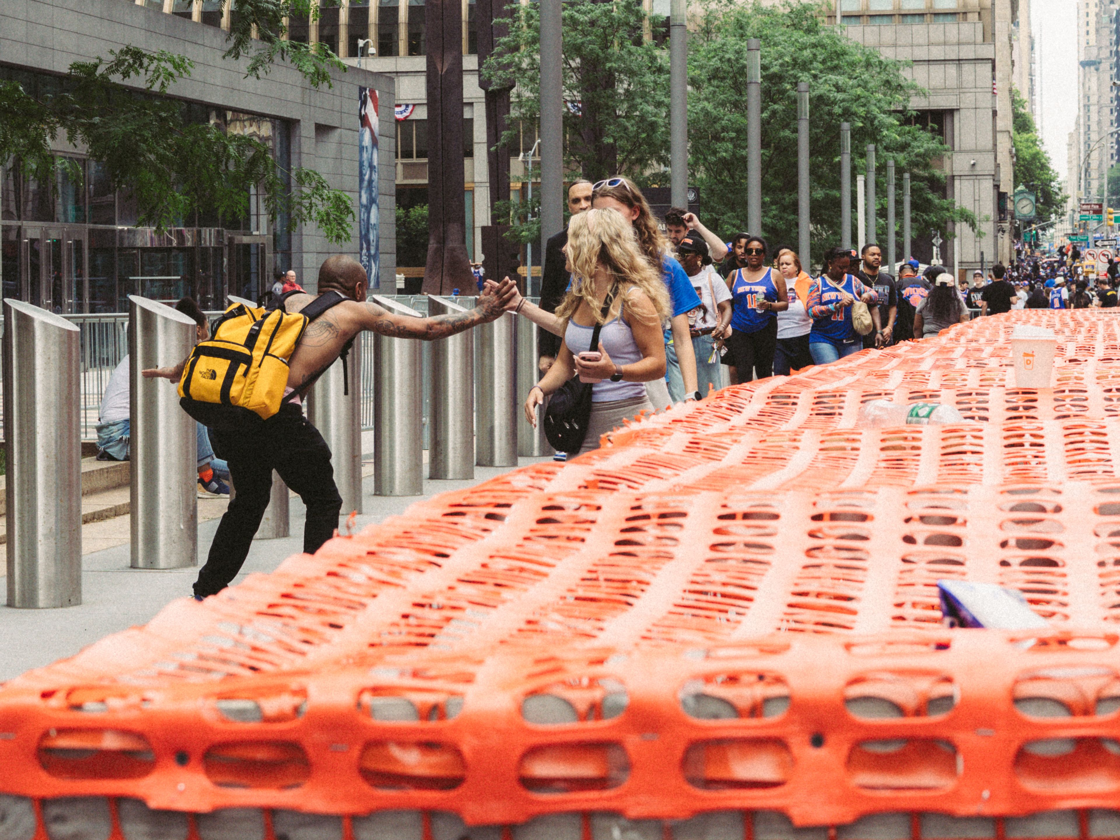 People greet each other during a lively urban parade, with an orange barrier and bustling street scene.