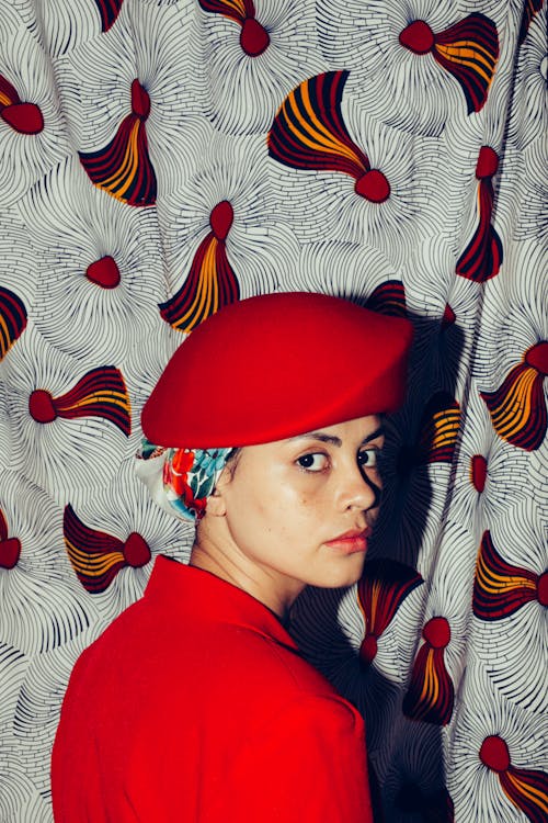 Back view of unemotional ethnic woman in retro hat and trendy jacket looking over shoulder on background of colorful curtain