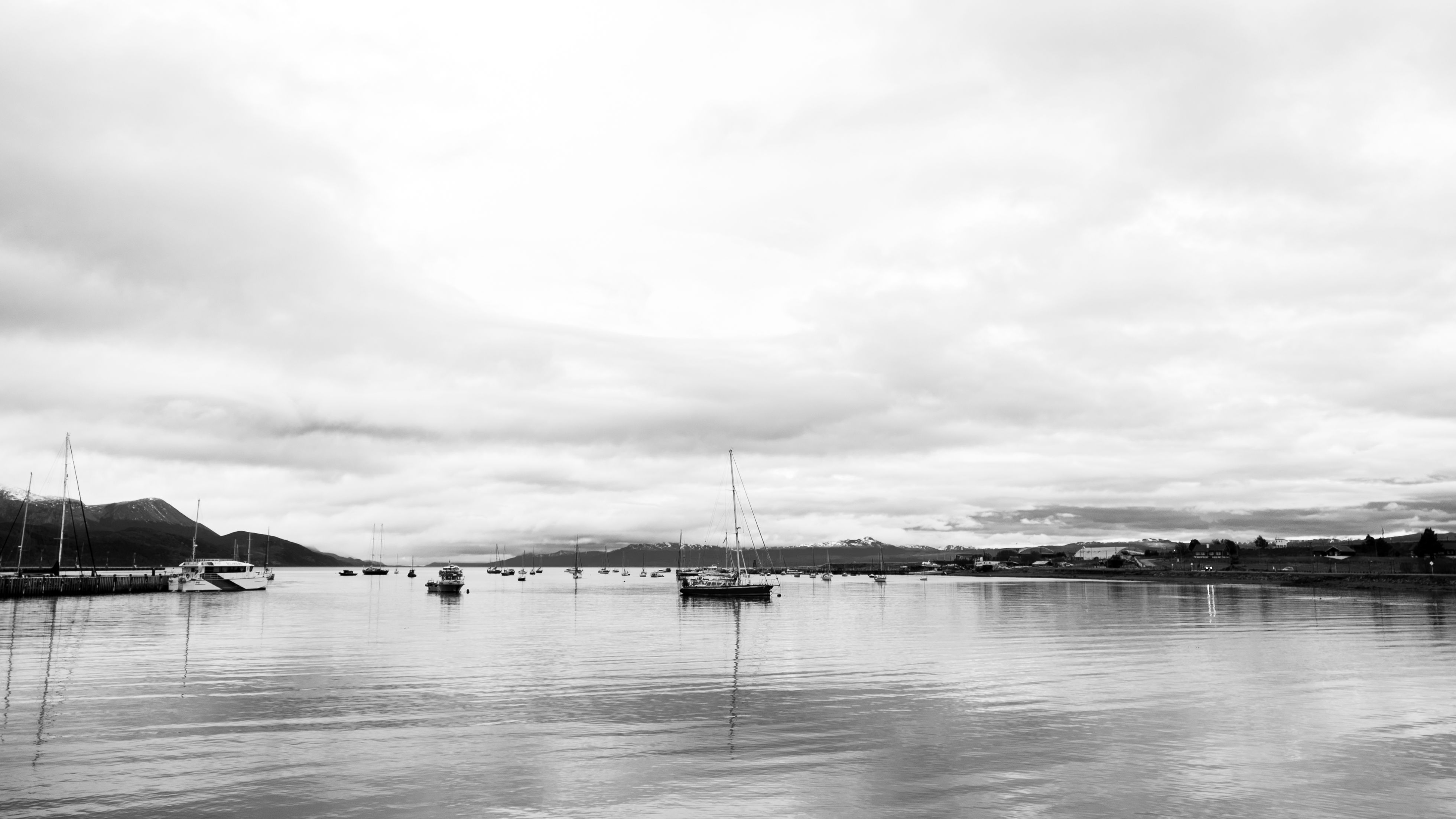 Black and white photo of Ushuaia harbor with sailboats and distant mountains.