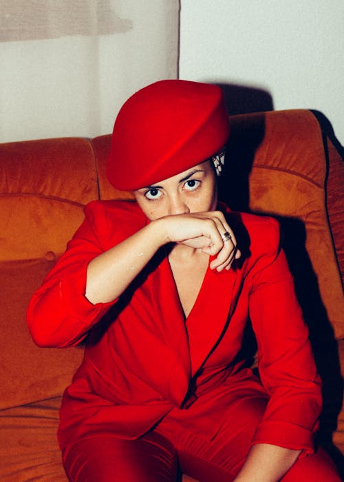 From above of determined female model wearing trendy suit and retro hat sitting in comfortable armchair and covering mouth with hand while looking at camera
