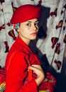 Side view of gorgeous woman wearing vintage hat and red costume standing in cozy living room with old fashioned handbag and looking at camera