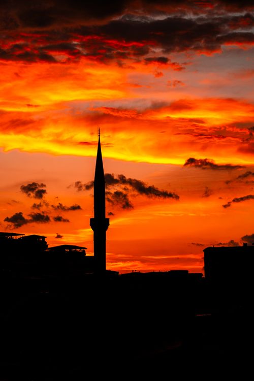 Free Silhouette of a minaret against a vivid orange sunset sky, creating a dramatic and striking visual scene. Stock Photo