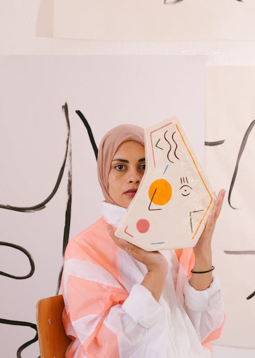 Pensive ethnic female wearing trendy clothes sitting on wooden chair with ceramic vase decorated with abstract pictures and looking at camera