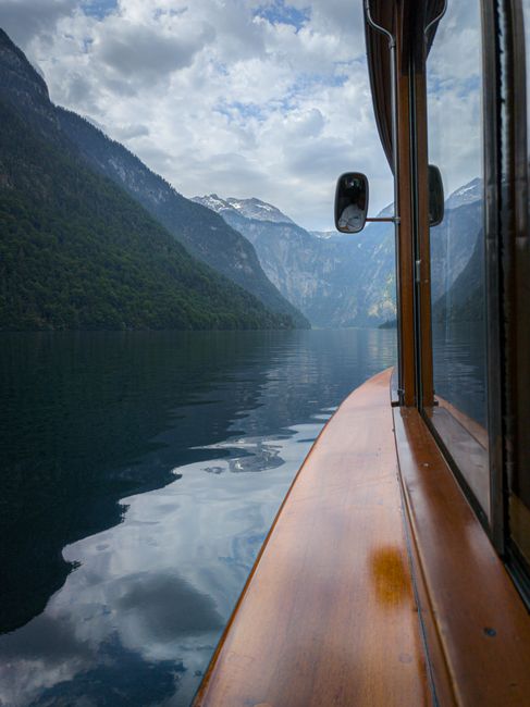 Serene view from a wooden boat on a tranquil mountain lake, surrounded by majestic peaks under cloudy skies.