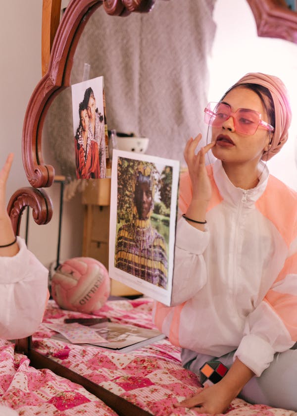 Young stylish female in eyeglasses and turban sitting on bed in bedroom and adjusting makeup while looking in mirror