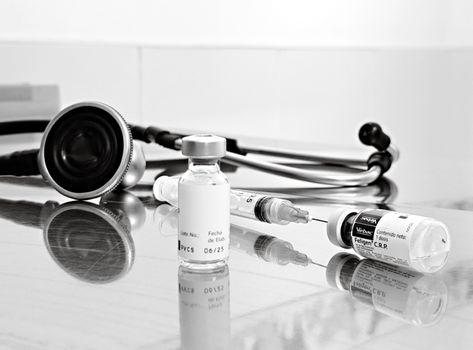 Black and white image of medical stethoscope next to vials and syringe on reflective surface.