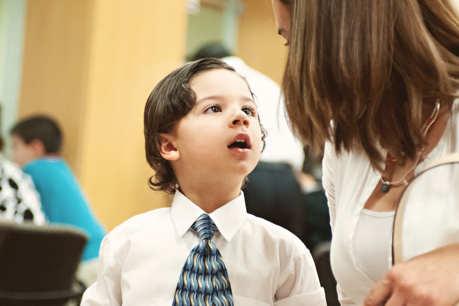 A young boy dressed formally in a tie, engaged in conversation with an adult indoors.