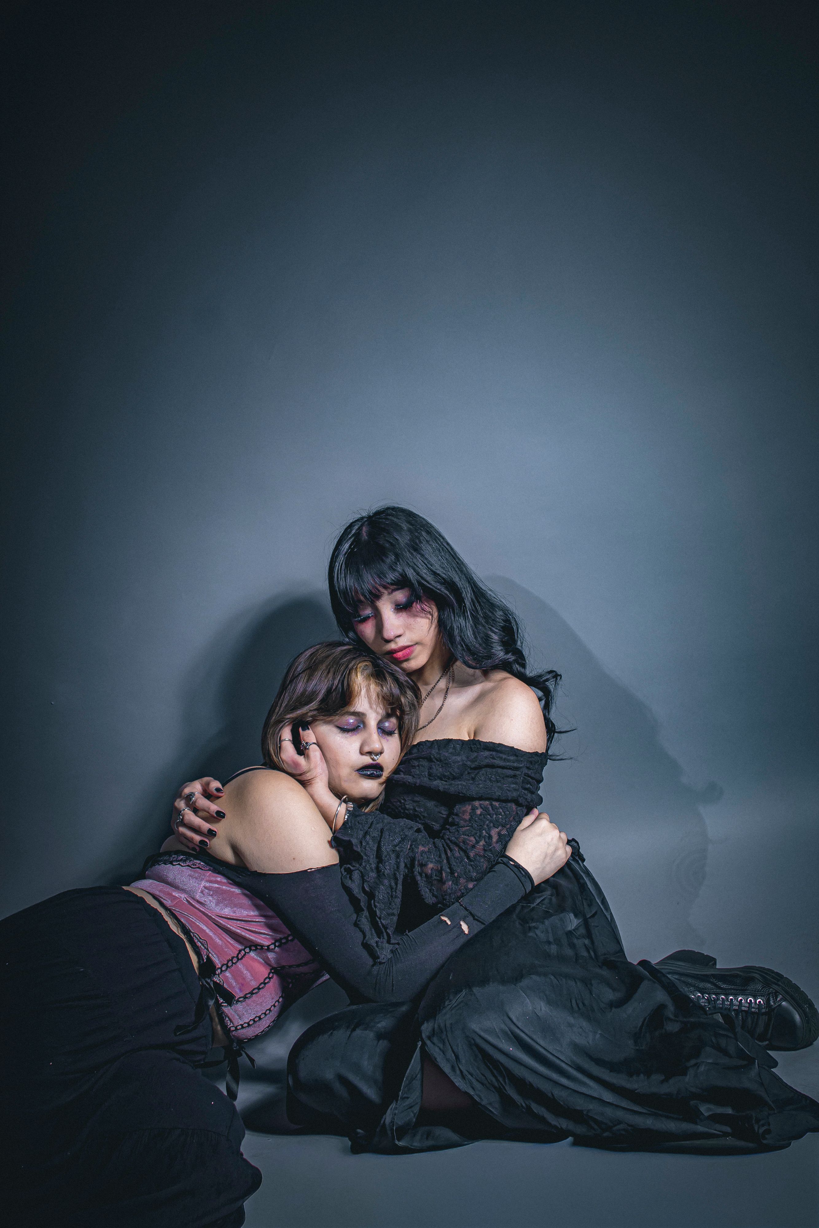 Two adults in gothic attire share a comforting embrace in a dimly lit setting.