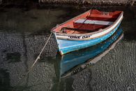Blue and red rowboat named 'One Day' tethered on calm reflective water, creating a tranquil scene.