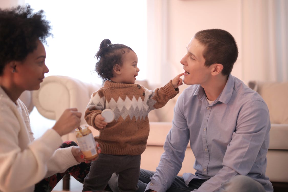 Side view of diverse parents in casual clothing sitting on floor and having fun with smiling toddler while gathering in cozy living room and having rest together
