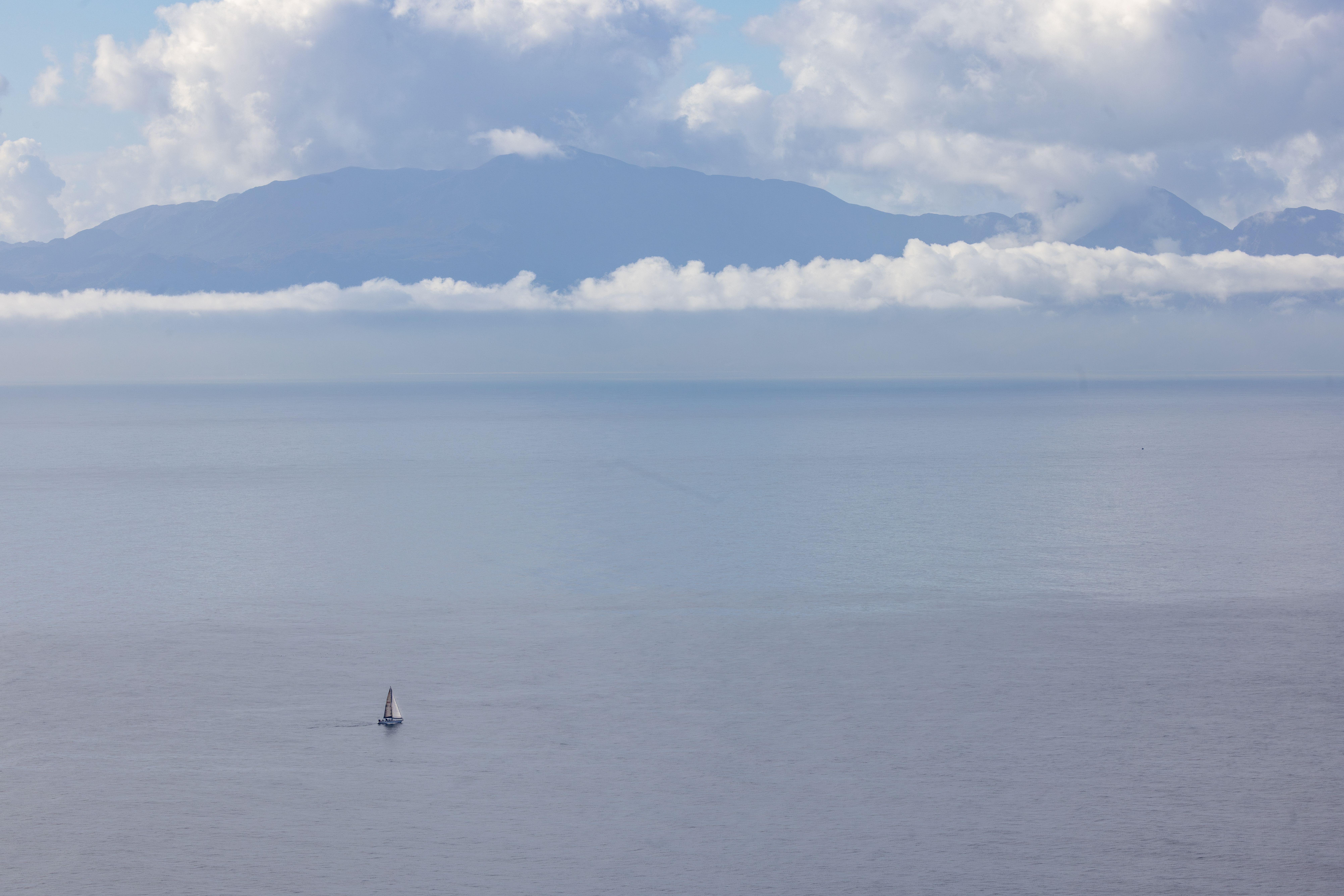 A serene view of a solitary sailboat on calm waters beneath a cloudy sky and distant mountains.