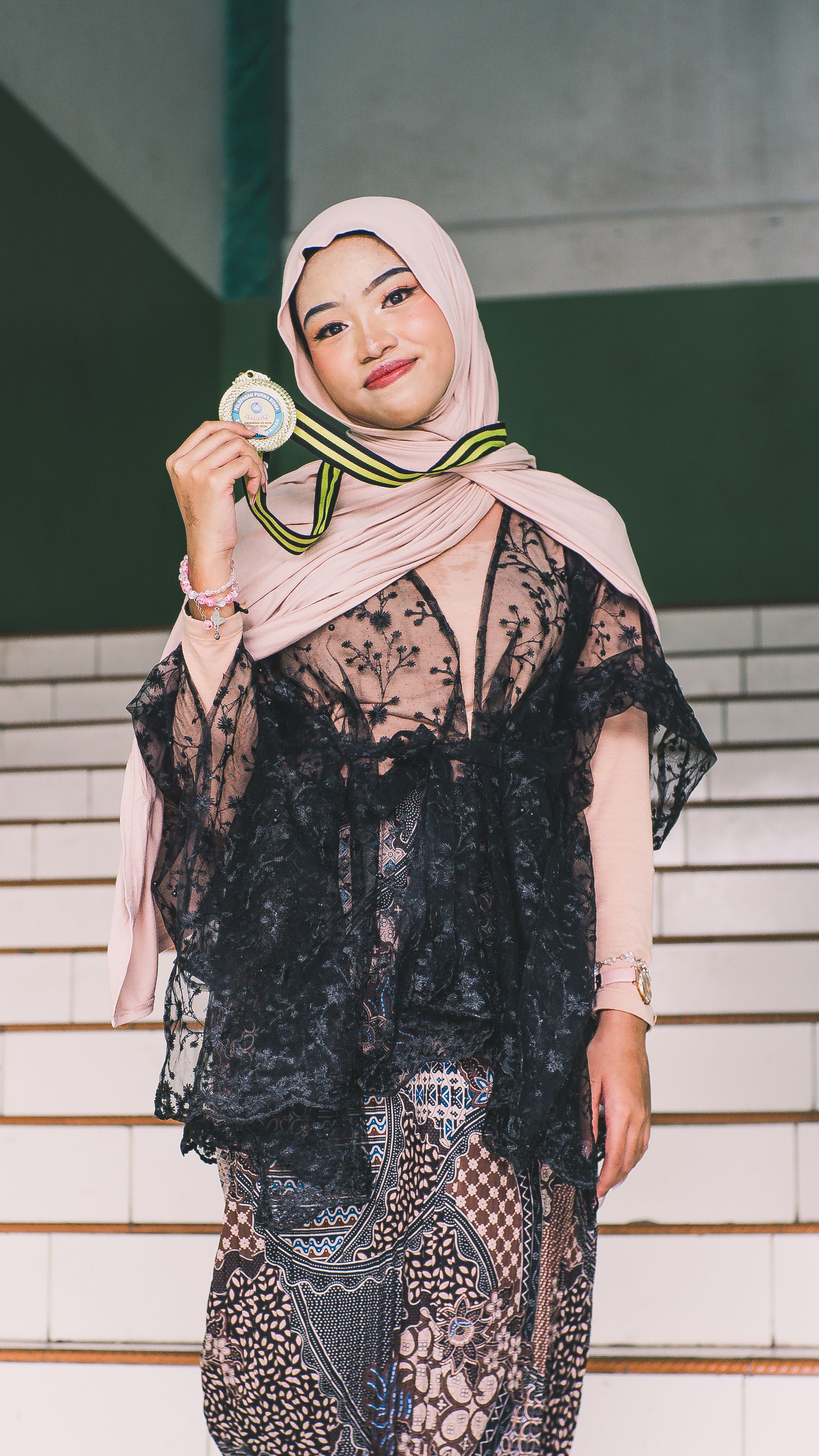 Elegant young woman wearing a hijab and holding a medal, posing confidently on a staircase.