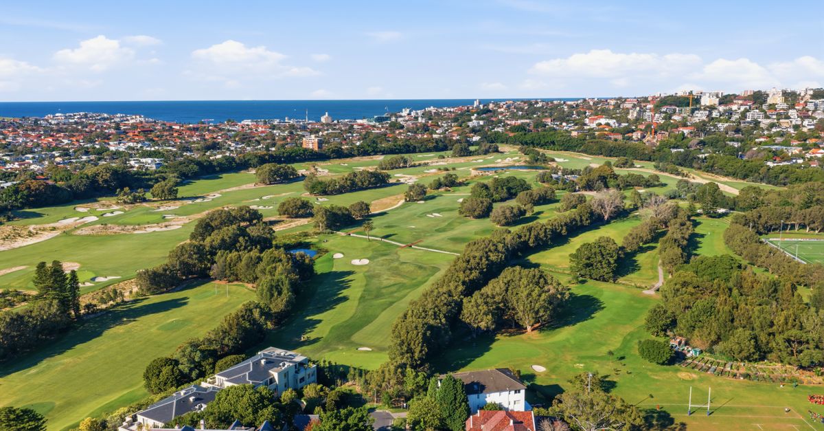 Aerial view of a golf course by the sea with cityscape in the background.