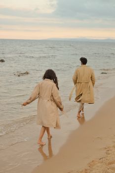 A couple strolls along a sandy beach at sunset, wearing beige trench coats.