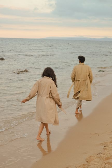 A couple strolls along a sandy beach at sunset, wearing beige trench coats.