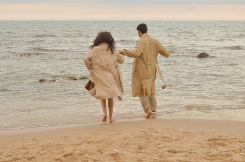 Free A romantic moment of a couple walking barefoot on the beach in trench coats. Stock Photo