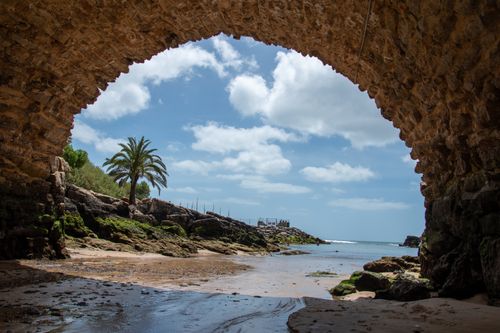 Free Beautiful coastal scene through a stone arch in Cascais, Portugal with a clear blue sky and palm tree. Stock Photo