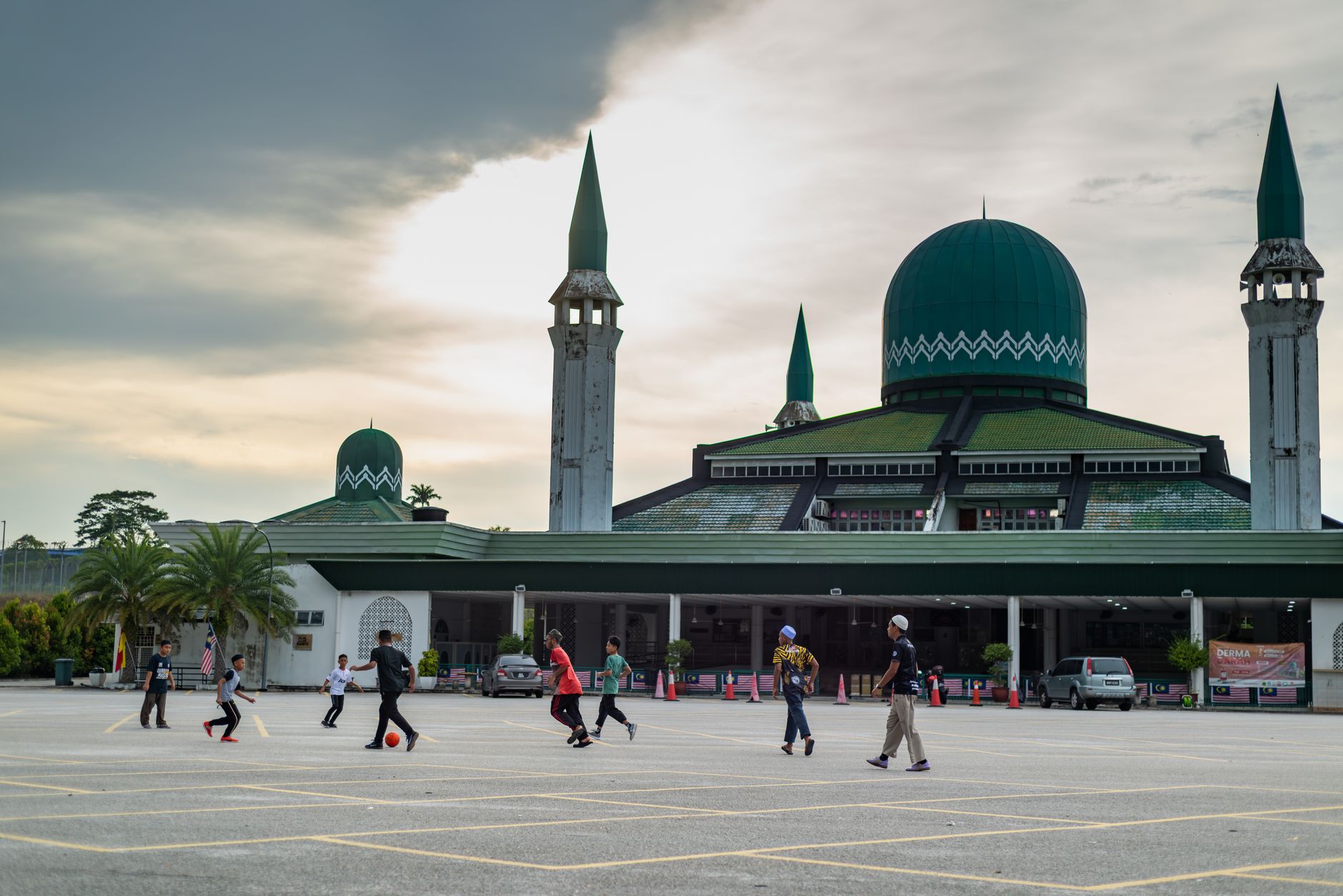 Kids play soccer in front of a mosque with green domes and minarets, under a cloudy sky.