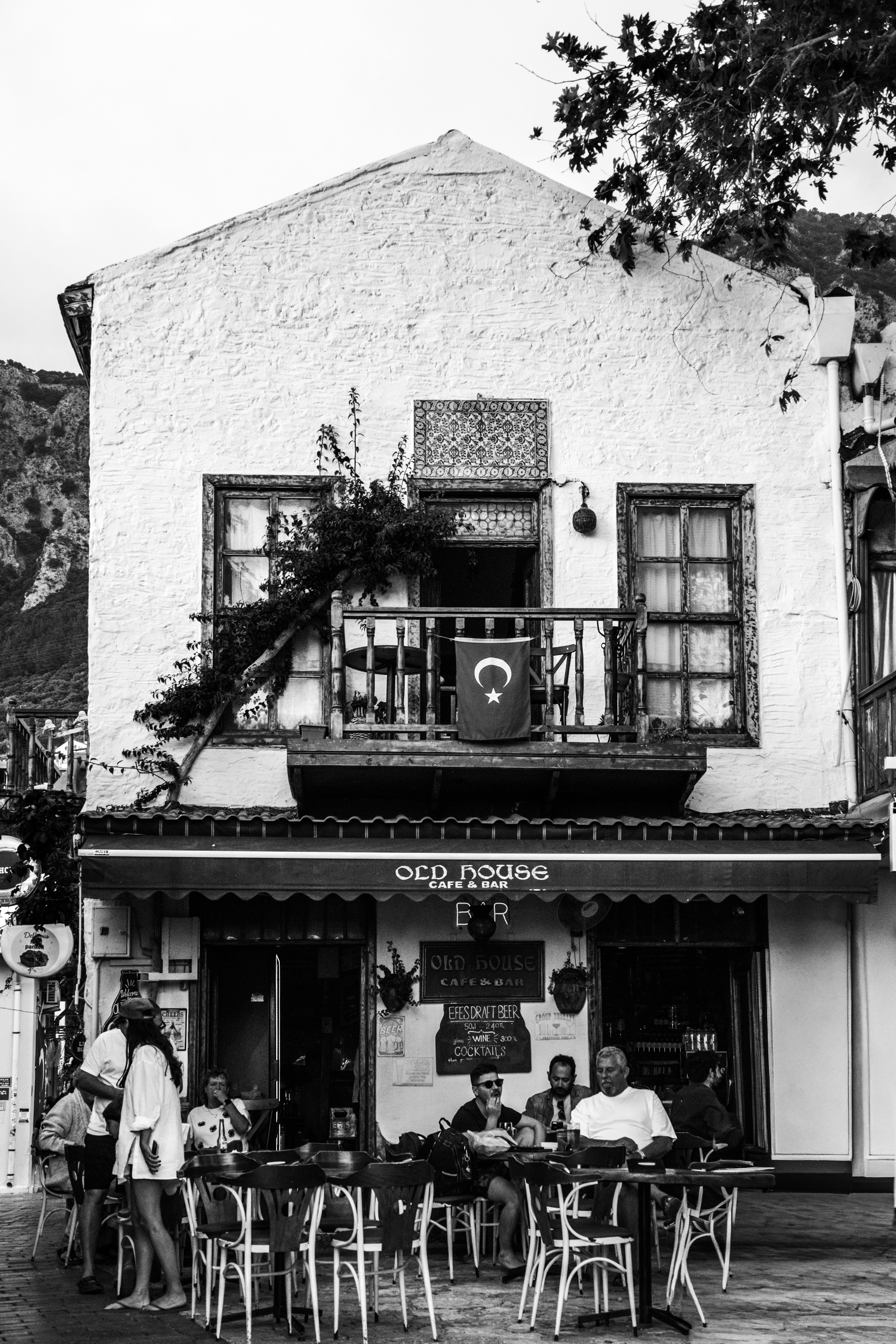Black and white photo of a charming café exterior with people sitting outdoors, Turkish flag visible.