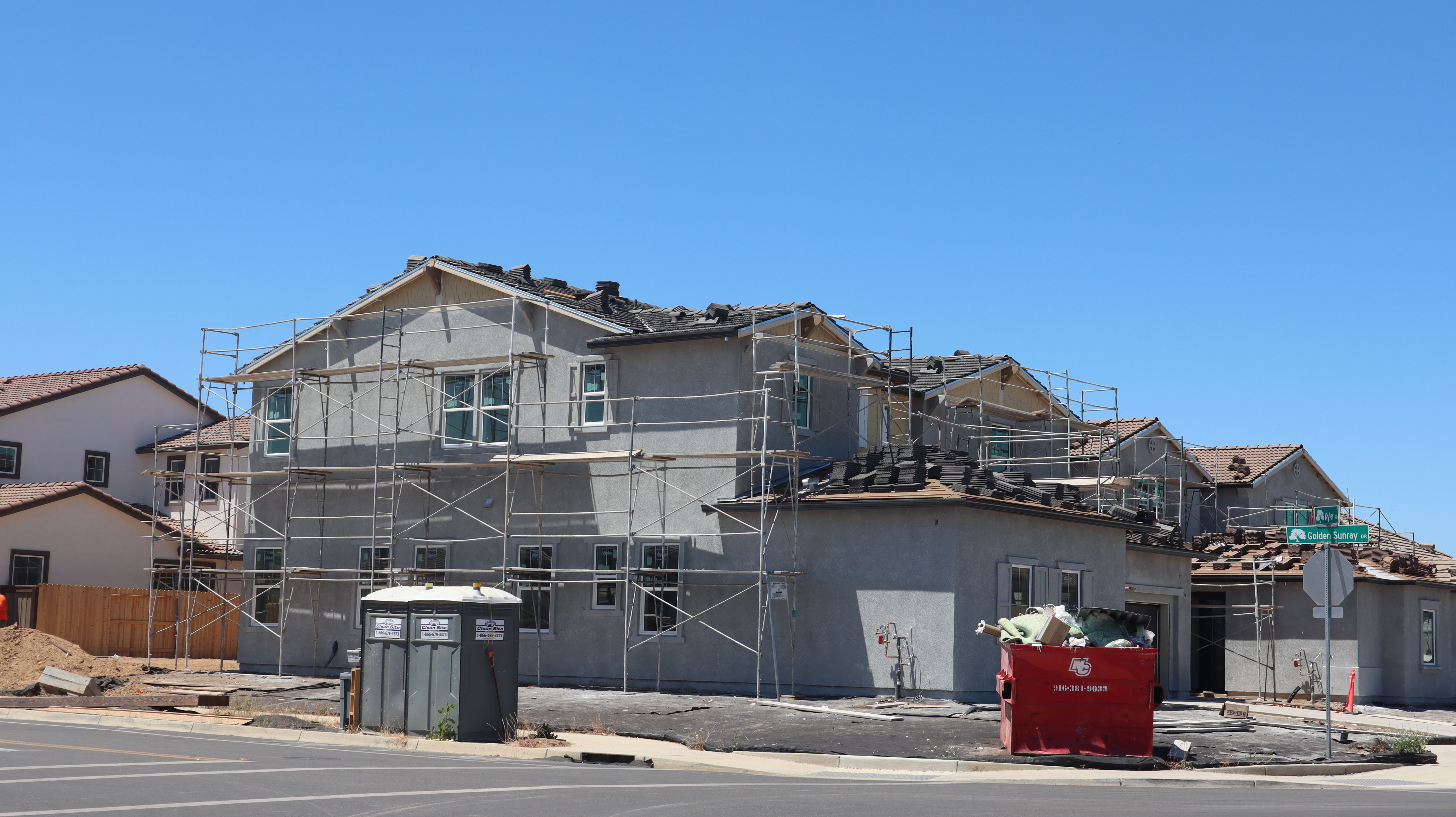 A row of modern suburban houses in various stages of construction under a clear blue sky, displaying exposed wood framing and protective wraps.