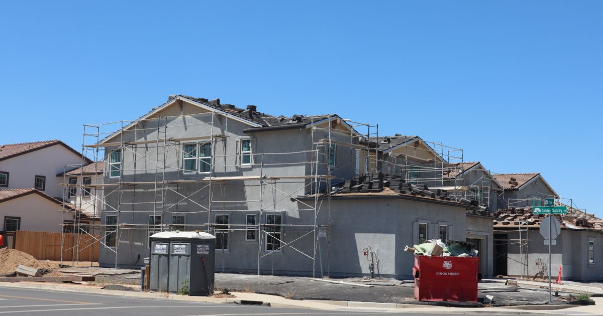 New two-story homes under construction on a California street, wrapped in scaffolding with tile roofs going on under a clear blue sky.