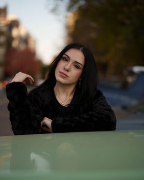 Portrait of a young woman in fashionable attire leaning on a car outdoors.