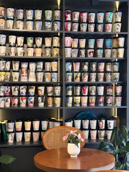 A vivid display of artistic coffee cups adorns the wall of a stylish café with a wooden table and plant.